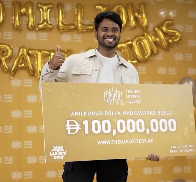 A man with short black hair and a smiling expression stands in front of a yellow wall decorated with large golden balloon letters spelling LUCKY DAY and gold balloons. He wears a light-colored blazer over a white shirt and dark pants. He holds a large golden rectangular check with black text reading THE UAE LOTTERY ANILKUMAR BOLLA MADHAVARAO BOLLA D100,000,000 at the bottom www.theuaelottery.ae DARE TO DREAM LUCKY DAY OCTOBER 2023. He gives a thumbs-up gesture with his right hand.