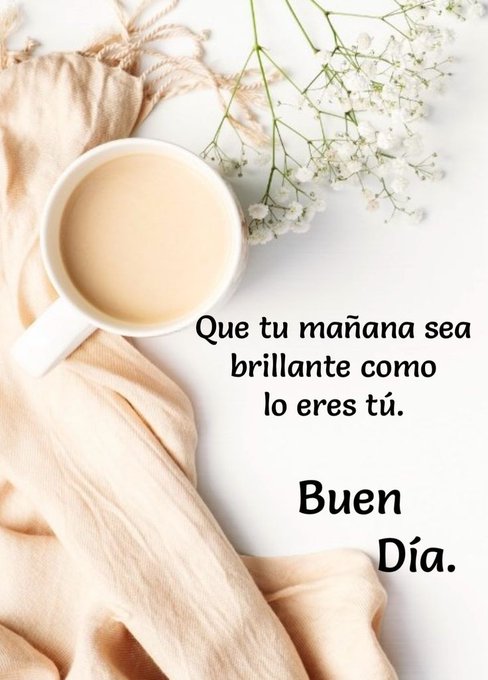 A white ceramic cup filled with light beige tea sits on a plain white surface. Surrounding the cup are folds of soft beige fabric draped nearby. Delicate white flowers with green stems are scattered around the cup and fabric. Overlaid text in black reads Que tu mañana sea brillante como lo eres tu at the top and Buen dia at the bottom.