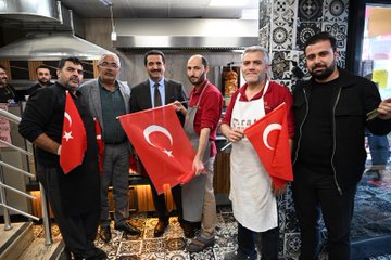 First image shows a group of men in suits and casual clothes standing on a paved area near buildings and trees, several holding red Turkish flags with white crescent and star, one elderly man with a cane and cap interacting with a suited man, others in vests and shirts nearby with bags. Second image depicts three men in suits and jackets on a walkway beside a grassy area with chairs and tables, each holding Turkish flags, Turkish flag banners hanging from poles and buildings in background under sunny sky. Third image features a diverse group of men and women in various outfits including headscarves and vests standing in shaded outdoor spot near restaurant buildings and tents, many holding Turkish flags, some smiling and posing together. Fourth image captures men in suits, aprons, and casual wear inside or near a kitchen area with grills and counters, holding Turkish flags, one preparing food with skewers, decorative tiles and lighting in background.