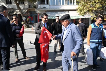 First image shows a group of men in suits and casual clothes standing on a paved area near buildings and trees, several holding red Turkish flags with white crescent and star, one elderly man with a cane and cap interacting with a suited man, others in vests and shirts nearby with bags. Second image depicts three men in suits and jackets on a walkway beside a grassy area with chairs and tables, each holding Turkish flags, Turkish flag banners hanging from poles and buildings in background under sunny sky. Third image features a diverse group of men and women in various outfits including headscarves and vests standing in shaded outdoor spot near restaurant buildings and tents, many holding Turkish flags, some smiling and posing together. Fourth image captures men in suits, aprons, and casual wear inside or near a kitchen area with grills and counters, holding Turkish flags, one preparing food with skewers, decorative tiles and lighting in background.