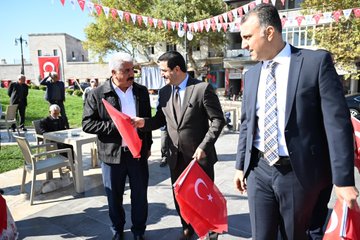 First image shows a group of men in suits and casual clothes standing on a paved area near buildings and trees, several holding red Turkish flags with white crescent and star, one elderly man with a cane and cap interacting with a suited man, others in vests and shirts nearby with bags. Second image depicts three men in suits and jackets on a walkway beside a grassy area with chairs and tables, each holding Turkish flags, Turkish flag banners hanging from poles and buildings in background under sunny sky. Third image features a diverse group of men and women in various outfits including headscarves and vests standing in shaded outdoor spot near restaurant buildings and tents, many holding Turkish flags, some smiling and posing together. Fourth image captures men in suits, aprons, and casual wear inside or near a kitchen area with grills and counters, holding Turkish flags, one preparing food with skewers, decorative tiles and lighting in background.