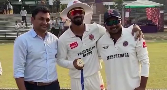 Three men stand on a grassy cricket field near stands and tents, one in a white shirt and trousers, the other two in white Maharashtra cricket uniforms with Punjab sponsor logos, one wearing a white hat with sunglasses, the other a cap with sunglasses, all smiling and posing with arms around each other, the middle man holding a trophy, background includes spectators and green awnings.