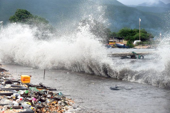 Large waves crash against a coastal area with debris scattered on the shore including plastic waste and a yellow container, boats are visible in the water and docked nearby, green trees and mountains in the background under a cloudy sky, a light pole stands near the scene.