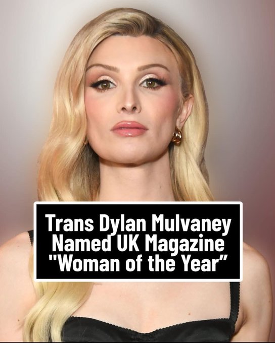 A woman with long wavy blonde hair, green eyes, neutral expression, wearing gold hoop earrings and pink lipstick, against a light gray background. Overlaid text box reads Trans Dylan Mulvaney Named UK Magazine Woman of the Year.