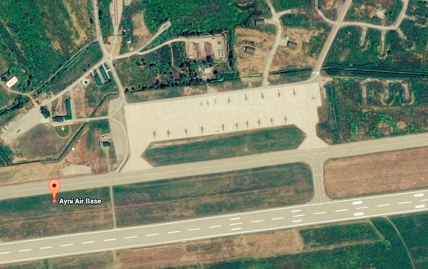 Satellite image displays the Ayni Air Base in Tajikistan with a red pin marker labeling it, showing a long runway, multiple aircraft parked on tarmac areas, surrounding buildings, roads, green fields, and sparse vegetation in a rural setting.