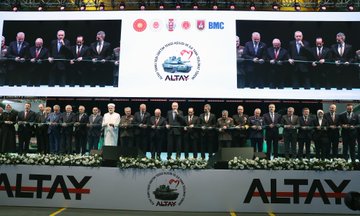 First image shows a man in dark suit and blue tie standing at a white podium with red Turkish emblem, speaking into microphones, with green military tank in background and barriers. Second image depicts the same man in suit standing beside large green armored vehicle labeled Altay in industrial hall with overhead cranes and Turkish flag emblem on wall. Third image features group of men in suits and one in yellow helmet with blue uniform examining interior of green machinery platform in factory setting with railings and equipment. Fourth image displays line of suited men including one in white headwear cutting ribbon at event stage with screens showing leaders, Altay logos, and floral decorations.