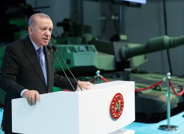 First image shows a man in dark suit and blue tie standing at a white podium with red Turkish emblem, speaking into microphones, with green military tank in background and barriers. Second image depicts the same man in suit standing beside large green armored vehicle labeled Altay in industrial hall with overhead cranes and Turkish flag emblem on wall. Third image features group of men in suits and one in yellow helmet with blue uniform examining interior of green machinery platform in factory setting with railings and equipment. Fourth image displays line of suited men including one in white headwear cutting ribbon at event stage with screens showing leaders, Altay logos, and floral decorations.