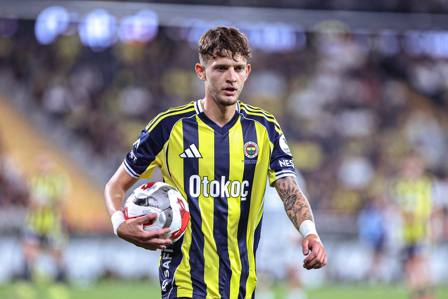 A young man with curly brown hair and a beard wearing a yellow and navy striped Fenerbahce soccer jersey with sponsor logos including OTOKOÇ holds a white and red soccer ball in his right hand while standing on a green grass field during a match with a blurred stadium crowd and lights in the background his left arm extended and yellow tattoo visible on his forearm.
