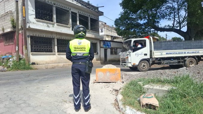 A police officer in a yellow high-visibility vest with PMT Villa Nueva text on the back stands facing away near a white dump truck loaded with dirt. The truck has a yellow light bar and is parked on a gravel road beside orange construction barriers. A rundown two-story building with barred windows and pink walls is visible to the left under power lines and trees. The officer wears a green helmet black pants and carries gear on a belt. Scattered dirt piles and grass are around the scene.