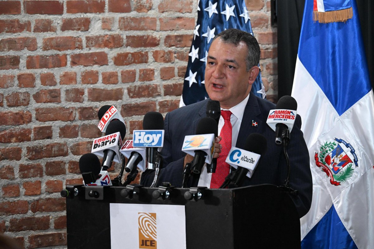 A man in a dark suit and red tie stands at a black podium with the JCE logo, speaking into multiple microphones labeled with media outlets like Hoy and CDN. Behind him are flags of the United States, Dominican Republic, and another country, against a brick wall. The setting appears to be a formal press conference.
