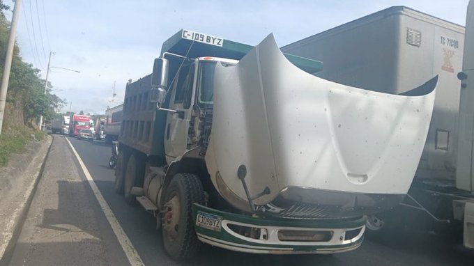 A green dump truck with license plate CJO8BYZ is positioned on the side of a road, its front hood lifted open revealing engine parts underneath. The truck appears damaged or under repair, with the white hood propped up. Surrounding vehicles including another truck and cars are visible on the busy roadway lined with utility poles and roadside vegetation under a clear sky.