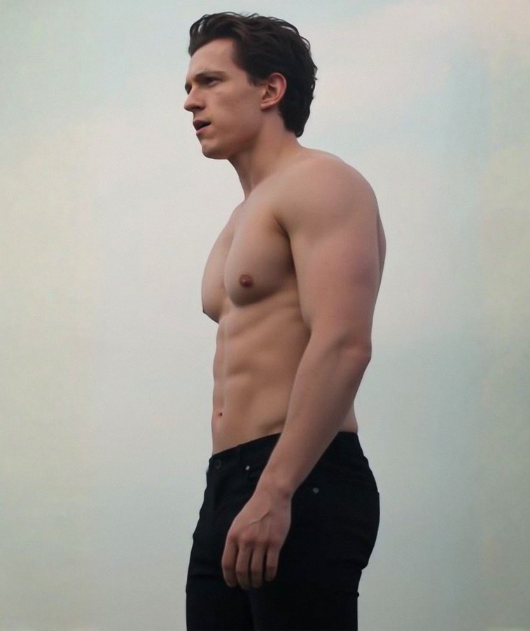 Tom Holland stands in profile view against a light blue-gray background, shirtless revealing defined chest and abdominal muscles, wearing dark jeans, with short dark hair and a neutral expression.