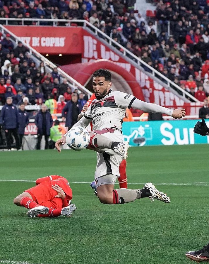 Soccer player in white and maroon uniform with long sleeves jumps and kicks a white soccer ball mid-air on green field. Another player in red uniform lies fallen on the ground below reaching out. Stadium stands filled with spectators in background. Red Copa de la Liga banner on upper structure. White sponsor logo on field side.
