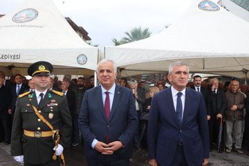 First image shows a group of people including suited officials and children gathered outside a building with Turkish flags, palm trees, and a police vehicle nearby. Second image depicts officials in suits and military uniforms standing under white tents with emblems, surrounded by attendees. Third image captures individuals holding flags in blue and red, with children and adults in casual and formal attire near a building facade. Fourth image features military personnel and officials saluting or standing formally outside a beige building with a tent.