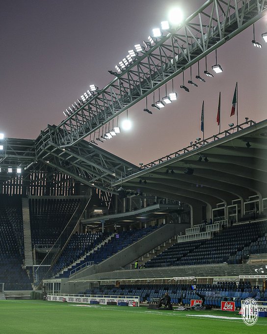 Nighttime view of a large football stadium with a vibrant green pitch in the foreground, surrounded by empty blue seating tiers and concrete structures. Bright white floodlights mounted on steel trusses overhead illuminate the field intensely against a deep purple twilight sky. Italian flags wave from poles near the stands, and advertising banners including one for a sponsor are visible along the pitch edges. The architecture features modern curved roofs and multiple levels of spectator areas, with no people or activity present on the field.