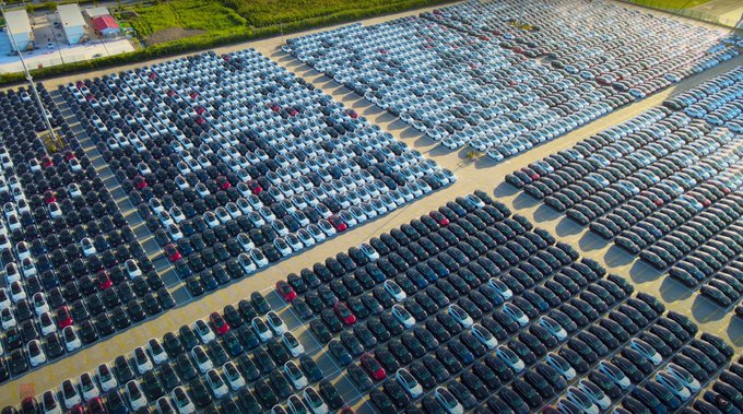 Aerial overhead view of a massive outdoor parking lot or delivery area filled with hundreds of Tesla electric vehicles in various colors including white, black, blue, and red, neatly arranged in rows on a paved surface, with surrounding green fields, a fence, and industrial buildings visible in the background under daylight conditions.