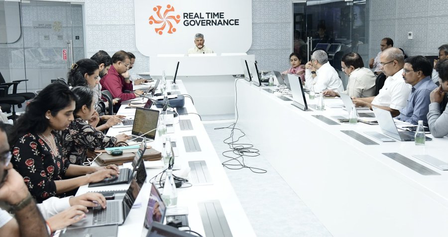 A group of officials including men and women in formal attire sit around a long white conference table with laptops and documents in a modern room featuring a podium and a backdrop displaying the Real Time Governance logo a circular orange design with swirling lines on a white panel behind the central seated figure at the podium.