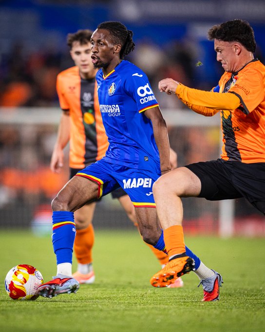 Soccer player in blue Getafe CF jersey with Mapelli sponsor and dreadlocks dribbles yellow ball on green field while orange-uniformed opponent in Villarreal kit extends arm to challenge, surrounded by teammates in similar kits and blurred stadium spectators.