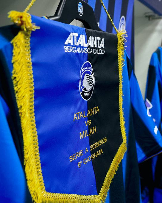 A black hanger suspends a rectangular banner in blue and black colors with yellow fringes along the edges. The banner displays the Atalanta BC emblem featuring a white siren figure on a blue background dated 1907. Text reads Atalanta Bergamo Calcio at the top. Below it states Atalanta vs Milan for Serie A 2022/2028 and 9 Giornata. The background shows a blurred locker room setting with additional hanging items.