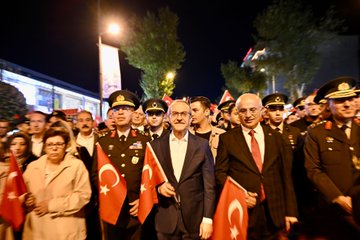 First image shows a group of officials and military personnel in formal uniforms and suits walking along a nighttime street lined with trees and lights many holding red Turkish flags with white crescent and star a man in suit and tie prominently in center with flag in hand women in coats and crowds in background illuminated by street lamps. Second image depicts a long nighttime street procession with numerous participants including women and men in casual clothes carrying a massive horizontal Turkish flag spanning the width of the road trees shops and banners visible along the sides under yellow streetlights. Third image captures a military marching band in dark green uniforms with gold accents playing brass instruments like trumpets and drums led by conductor with baton walking on a lit street at night surrounded by spectators and buildings. Fourth image features four men two in military uniforms with hats and two in suits standing at a wooden podium one speaking into a red microphone with wooden stand lit area behind with screens and flags in background nighttime urban setting.