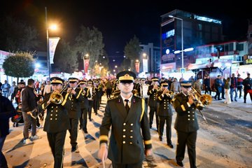 First image shows a group of officials and military personnel in formal uniforms and suits walking along a nighttime street lined with trees and lights many holding red Turkish flags with white crescent and star a man in suit and tie prominently in center with flag in hand women in coats and crowds in background illuminated by street lamps. Second image depicts a long nighttime street procession with numerous participants including women and men in casual clothes carrying a massive horizontal Turkish flag spanning the width of the road trees shops and banners visible along the sides under yellow streetlights. Third image captures a military marching band in dark green uniforms with gold accents playing brass instruments like trumpets and drums led by conductor with baton walking on a lit street at night surrounded by spectators and buildings. Fourth image features four men two in military uniforms with hats and two in suits standing at a wooden podium one speaking into a red microphone with wooden stand lit area behind with screens and flags in background nighttime urban setting.