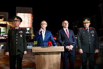 First image shows a group of officials and military personnel in formal uniforms and suits walking along a nighttime street lined with trees and lights many holding red Turkish flags with white crescent and star a man in suit and tie prominently in center with flag in hand women in coats and crowds in background illuminated by street lamps. Second image depicts a long nighttime street procession with numerous participants including women and men in casual clothes carrying a massive horizontal Turkish flag spanning the width of the road trees shops and banners visible along the sides under yellow streetlights. Third image captures a military marching band in dark green uniforms with gold accents playing brass instruments like trumpets and drums led by conductor with baton walking on a lit street at night surrounded by spectators and buildings. Fourth image features four men two in military uniforms with hats and two in suits standing at a wooden podium one speaking into a red microphone with wooden stand lit area behind with screens and flags in background nighttime urban setting.