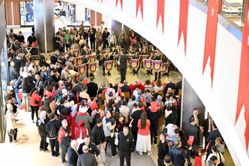 First image shows a group of children and adults in a brightly lit indoor mall area with FLO store signage, many holding and waving red Turkish flags with white crescent and star, a young girl in light clothing extending a flag towards seated officials including men in suits and military uniform smiling and engaging. Second image depicts a diverse crowd including women in headscarves and children in casual wear seated and standing in a Skechers store section, enthusiastically waving Turkish flags overhead during an event. Third image captures an overhead view of a large gathering in a curved mall atrium with red banners hanging from railings, a central group of performers in purple and gold uniforms playing instruments on a stage area surrounded by spectators in red attire holding flags. Fourth image features a close-up of several young girls in colorful sweaters and dresses holding Turkish flags, standing with elderly men in glasses and suits, all smiling and posing together in an indoor public space with other attendees in background.