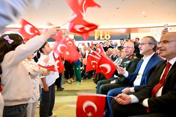 First image shows a group of children and adults in a brightly lit indoor mall area with FLO store signage, many holding and waving red Turkish flags with white crescent and star, a young girl in light clothing extending a flag towards seated officials including men in suits and military uniform smiling and engaging. Second image depicts a diverse crowd including women in headscarves and children in casual wear seated and standing in a Skechers store section, enthusiastically waving Turkish flags overhead during an event. Third image captures an overhead view of a large gathering in a curved mall atrium with red banners hanging from railings, a central group of performers in purple and gold uniforms playing instruments on a stage area surrounded by spectators in red attire holding flags. Fourth image features a close-up of several young girls in colorful sweaters and dresses holding Turkish flags, standing with elderly men in glasses and suits, all smiling and posing together in an indoor public space with other attendees in background.