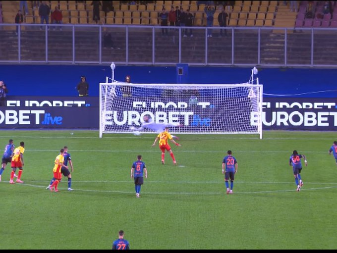 A soccer goal with white net and posts stands on a green field surrounded by blue barriers advertising Eurobet Live. Several players in red and blue jerseys are positioned near the goal, with one player in a yellow jersey diving to save a penalty kick from a player in a blue jersey approaching the ball. The stadium seating and barriers are visible in the background.