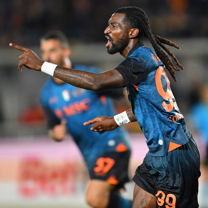 Victor Osimhen with long dreadlocks wearing a blue Napoli jersey numbered 9 points emphatically his expression intense and mouth open in celebration during a match another teammate in blue kit with number 97 runs nearby stadium stands with blurred crowd in background orange accents on shorts and wristbands visible.