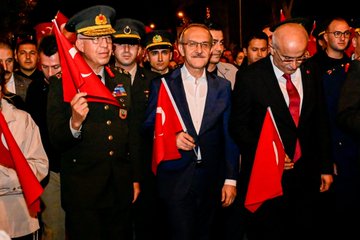 First image shows a group of men in military uniforms and suits holding Turkish flags during an evening outdoor event, with one central figure in glasses and suit carrying a flag, surrounded by officers in caps and formal attire under dim lighting. Second image depicts a diverse group including men in suits, a woman in headscarf, and police officers in vests inside a building with wooden paneling, some wearing caps with police emblem. Third image is an aerial night view of a large crowd of people in dark clothing holding red Turkish flags gathered around police vehicles with lights on a street lined with trees and buildings. Fourth image is an overhead night shot of a long curved line of illuminated orange crescent moon shapes along a street with trees, buildings, and scattered purple lights.