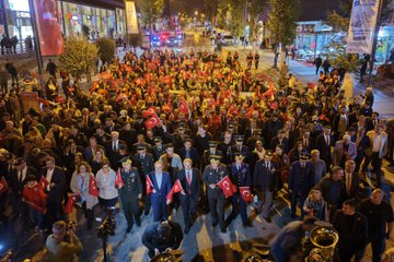 First image shows a group of men in military uniforms and suits holding Turkish flags during an evening outdoor event, with one central figure in glasses and suit carrying a flag, surrounded by officers in caps and formal attire under dim lighting. Second image depicts a diverse group including men in suits, a woman in headscarf, and police officers in vests inside a building with wooden paneling, some wearing caps with police emblem. Third image is an aerial night view of a large crowd of people in dark clothing holding red Turkish flags gathered around police vehicles with lights on a street lined with trees and buildings. Fourth image is an overhead night shot of a long curved line of illuminated orange crescent moon shapes along a street with trees, buildings, and scattered purple lights.