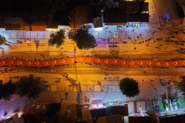 First image shows a group of men in military uniforms and suits holding Turkish flags during an evening outdoor event, with one central figure in glasses and suit carrying a flag, surrounded by officers in caps and formal attire under dim lighting. Second image depicts a diverse group including men in suits, a woman in headscarf, and police officers in vests inside a building with wooden paneling, some wearing caps with police emblem. Third image is an aerial night view of a large crowd of people in dark clothing holding red Turkish flags gathered around police vehicles with lights on a street lined with trees and buildings. Fourth image is an overhead night shot of a long curved line of illuminated orange crescent moon shapes along a street with trees, buildings, and scattered purple lights.
