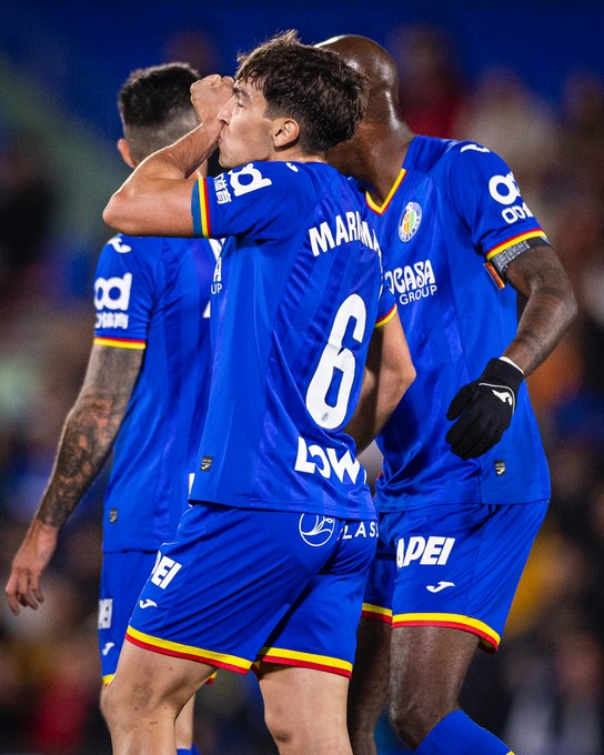 Three male soccer players in blue Getafe CF jerseys with red and yellow accents stand on a green field embracing each other in celebration one player has number 6 on back and Mahou sponsor logo another wears black gloves and Las Delicias sponsor on shorts stadium seating visible in background.