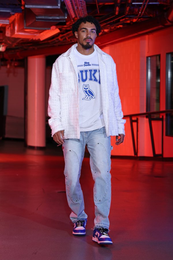 Young Black male with curly hair and mustache stands in indoor arena under red lighting and high ceilings with exposed beams. He wears open white jacket displaying Duke text and blue owl logo over white t-shirt, blue jeans with rips at knees, and purple low-top Nike sneakers with white soles. His expression is neutral as he walks forward.