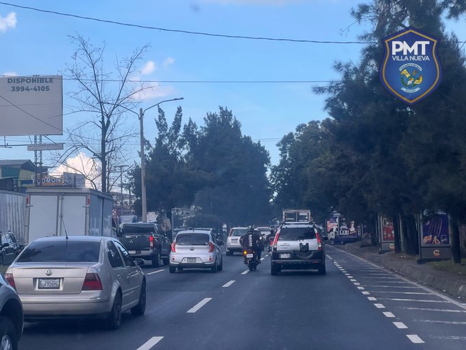 Street scene with multiple cars including sedans and SUVs lined up on a multi-lane road under partly cloudy sky. Trees and utility poles line the roadside. A police vehicle with PMT VILLA NUEVA emblem is visible among the traffic. A billboard displays numbers 299-410. Motorcycles and pedestrians are absent in the immediate view.