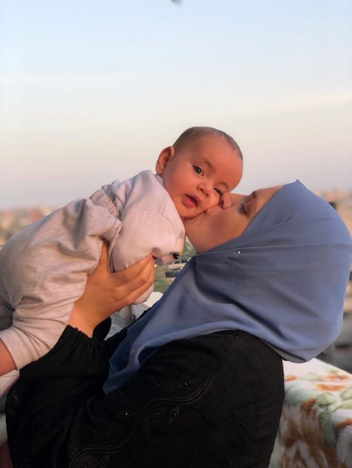 A woman wearing a blue hijab and black clothing holds and kisses a baby boy in white outfit on her cheek, with the babys face showing curiosity, set against a clear sky with buildings in the distant background.