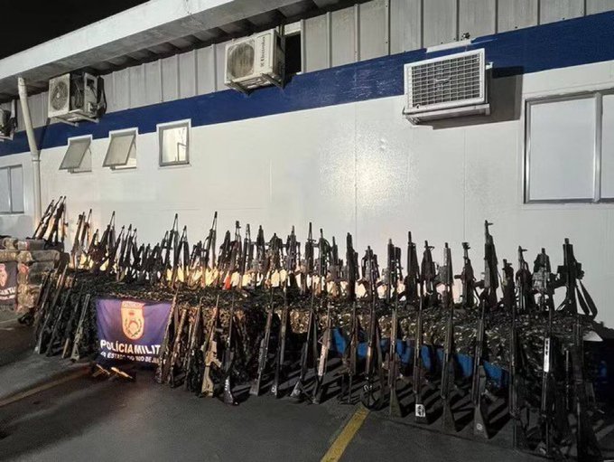 Numerous assault rifles arranged in rows on the ground in front of a police banner reading Policia Militar, displayed outdoors near a building with air conditioning units and windows under nighttime conditions.