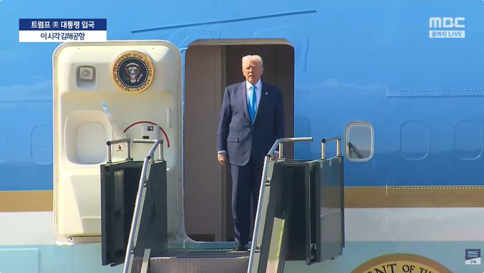 Blue airplane with open door and stairs, white structure with gold US presidential seal beside the entrance, Donald Trump in dark suit and blue tie standing at the doorway looking outward, blue MBC broadcast overlay with Korean text indicating live coverage from the US.