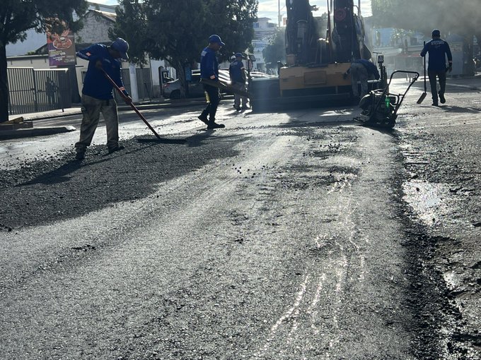 First image shows several workers in blue uniforms and helmets engaged in road paving on a sunny street lined with trees and buildings using a large yellow asphalt roller machine a green blower device and brooms to smooth fresh black asphalt over the road surface with some workers walking nearby. Second image depicts a busy urban street intersection with green and white traffic cones placed around a construction zone several motorcycles including orange and black ones parked or moving near the cones a black SUV and a white scooter with riders wearing helmets and buildings with signs like EL MUNDO in the background under clear daylight.