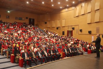 First image shows a large auditorium filled with seated audience members in formal attire including suits and ties, viewed from the stage with red chairs and wooden paneling. Second image depicts a man in a dark suit with tie speaking into a microphone on stage, standing near banners for educational organizations and a screen. Third image captures the auditorium with audience seated and a man in suit speaking on stage from a side angle. Fourth image features two men in suits holding a green ornate plaque on stage, with a projection screen displaying event title Türkiye Yüzyılı Maarif Modeli and names Prof. Dr. Cihad Demirli and others.