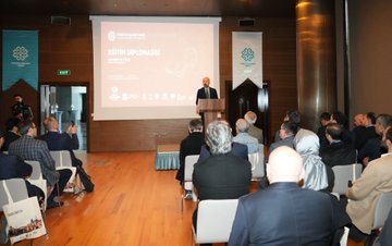 A bald man in a dark suit and tie stands at a wooden podium gesturing with both hands while speaking, with a projection screen behind showing abstract designs and logos including Turkiye Maarif Vakfi, in a modern conference room with wooden panels and a white banner. Several attendees including men and women in formal attire sit in rows of chairs facing the stage, some taking notes, in a conference hall with green and white banners and a screen displaying event details. A man in a suit speaks at a podium in front of a screen showing text about the event, with an audience of diverse individuals in business casual and traditional attire seated and listening attentively. A group of diverse attendees including women in headscarves and men in suits listen to a speaker holding a microphone, seated in chairs arranged in rows in a conference setting with wooden floors and modern decor.