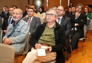 A bald man in a dark suit and tie stands at a wooden podium gesturing with both hands while speaking, with a projection screen behind showing abstract designs and logos including Turkiye Maarif Vakfi, in a modern conference room with wooden panels and a white banner. Several attendees including men and women in formal attire sit in rows of chairs facing the stage, some taking notes, in a conference hall with green and white banners and a screen displaying event details. A man in a suit speaks at a podium in front of a screen showing text about the event, with an audience of diverse individuals in business casual and traditional attire seated and listening attentively. A group of diverse attendees including women in headscarves and men in suits listen to a speaker holding a microphone, seated in chairs arranged in rows in a conference setting with wooden floors and modern decor.