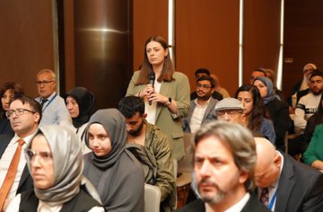 A bald man in a dark suit and tie stands at a wooden podium gesturing with both hands while speaking, with a projection screen behind showing abstract designs and logos including Turkiye Maarif Vakfi, in a modern conference room with wooden panels and a white banner. Several attendees including men and women in formal attire sit in rows of chairs facing the stage, some taking notes, in a conference hall with green and white banners and a screen displaying event details. A man in a suit speaks at a podium in front of a screen showing text about the event, with an audience of diverse individuals in business casual and traditional attire seated and listening attentively. A group of diverse attendees including women in headscarves and men in suits listen to a speaker holding a microphone, seated in chairs arranged in rows in a conference setting with wooden floors and modern decor.