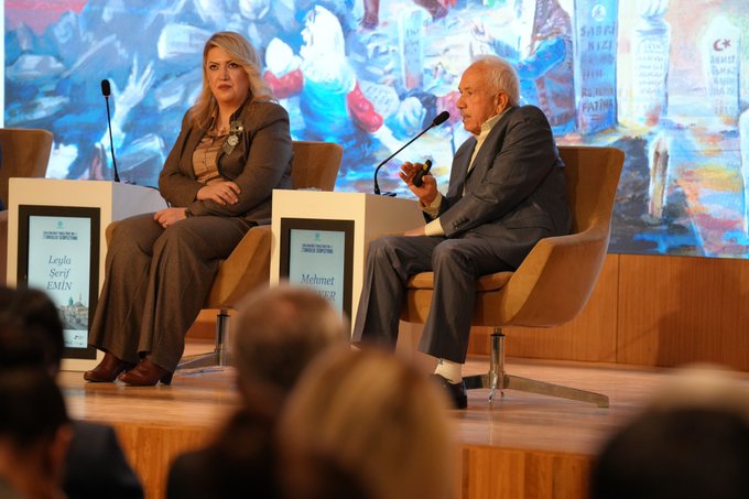 An elderly man with white hair and a mustache wearing a light blue shirt and dark gray suit sits in a beige armchair holding a microphone while speaking into another on a stand in front of a beige screen and a white statue. A second image shows the same elderly man in a dark gray suit sitting in a beige armchair holding a microphone next to a blonde woman in a brown suit and pants sitting in another armchair with two microphones on stands in front of beige podiums with text on screens and a colorful abstract background.