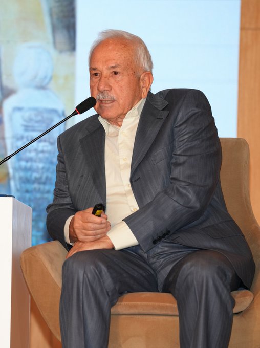 An elderly man with white hair and a mustache wearing a light blue shirt and dark gray suit sits in a beige armchair holding a microphone while speaking into another on a stand in front of a beige screen and a white statue. A second image shows the same elderly man in a dark gray suit sitting in a beige armchair holding a microphone next to a blonde woman in a brown suit and pants sitting in another armchair with two microphones on stands in front of beige podiums with text on screens and a colorful abstract background.