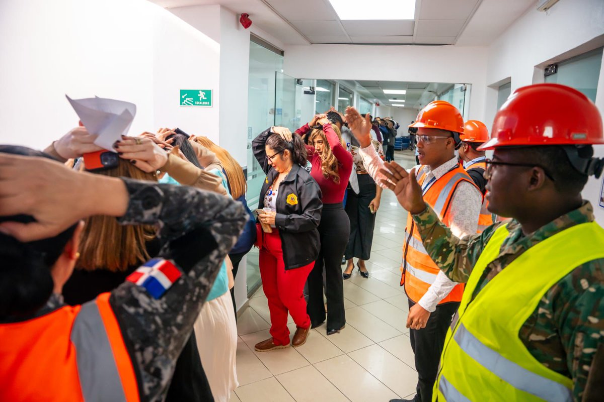 Group of people including women in casual clothing and men in orange safety vests and hard hats gathered in a modern hallway with white walls glass doors and emergency exit signs some holding papers and interacting near a Dominican Republic flag