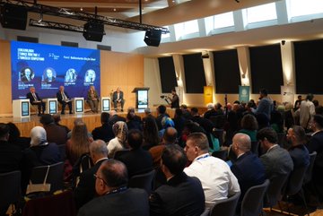 An elderly man with white hair and mustache in a dark suit and blue tie speaks into a microphone while seated, gesturing with his hand, against a backdrop with French text L'ancien Secrétaire général de la Présidence. A conference room with a large blue screen displaying Balkanlarda Göçün İzleri and photos of four panelists including Ahmet Balseven, Prof. Dr. Mustafa İsen, Leyla Şerif Emin, and Mehmet Türkeş, shows four individuals in formal attire seated at a panel table with microphones and screens, surrounded by an audience in business clothes. Close-up of the panel with the four speakers engaged in discussion, backdrop featuring their names and photos, audience visible in the foreground wearing varied attire including suits and headscarves.
