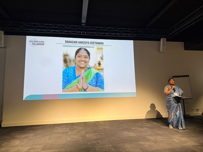 A woman in a blue saree stands on a stage holding a microphone and papers addressing an audience with a large projection screen behind her displaying the text Danasari Seethakka and an image of the same woman smiling with hands pressed together in greeting wearing a green saree against a light blue and white background in a conference room setting with beige walls speakers and a wooden floor.