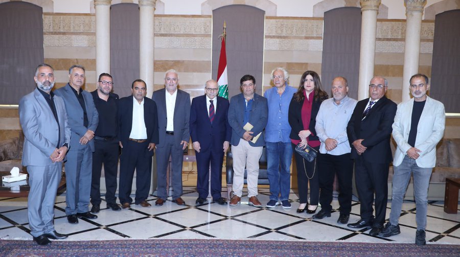 A group of eighteen formally dressed men and women stands in two rows in an ornate room with beige stone walls arched doorways draped flags including the Lebanese flag and a table with a white cloth in the foreground all posing seriously for a professional photograph.